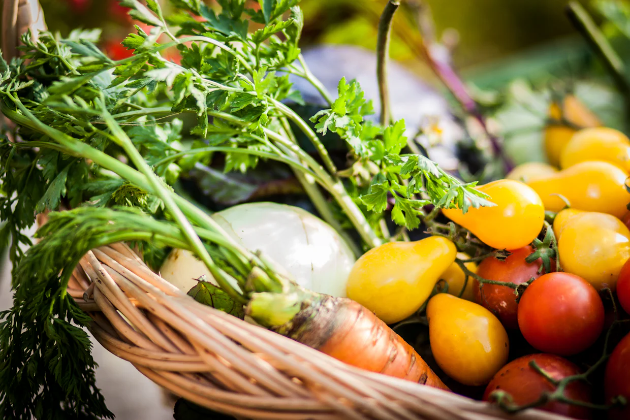 Un panier en osier rempli de légumes : tomates, carottes, oignons et verdure.