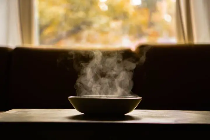 Un bol de soupe fumant sur une table en bois, près d'une fenêtre.