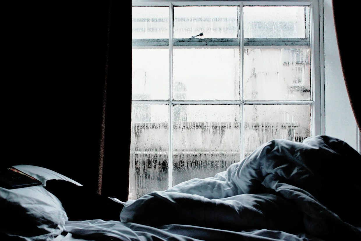 Un lit défait avec une couette froissée près d'une fenêtre couverte de condensation.