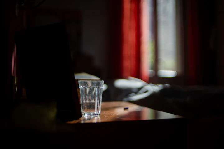 Un verre d'eau sur une table de chevet dans une pièce sombre, une fenêtre aux rideaux tirés derrière.