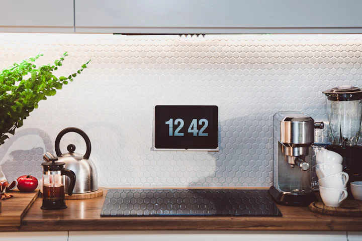 Un comptoir de cuisine à la lumière du jour avec une bouilloire, une cafetière à piston, une fougère et une horloge au mur.