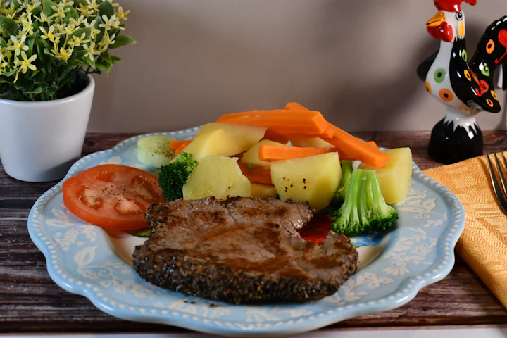 Une assiette de steak avec des pommes de terre bouillies, des carottes, du brocoli et une tranche de tomate sur une table de bois.