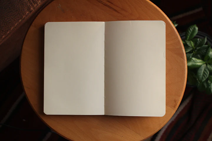 A plain notebook open on a wooden table with a pen resting on it.