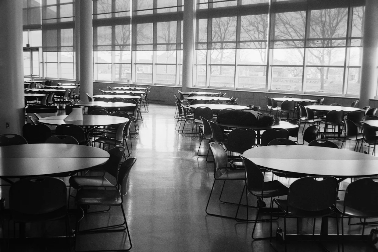 An empty cafeteria with rows of round tables and chairs by tall windows.