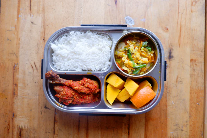 A packed lunch container with rice, chicken, vegetables and fruit on a wooden counter.