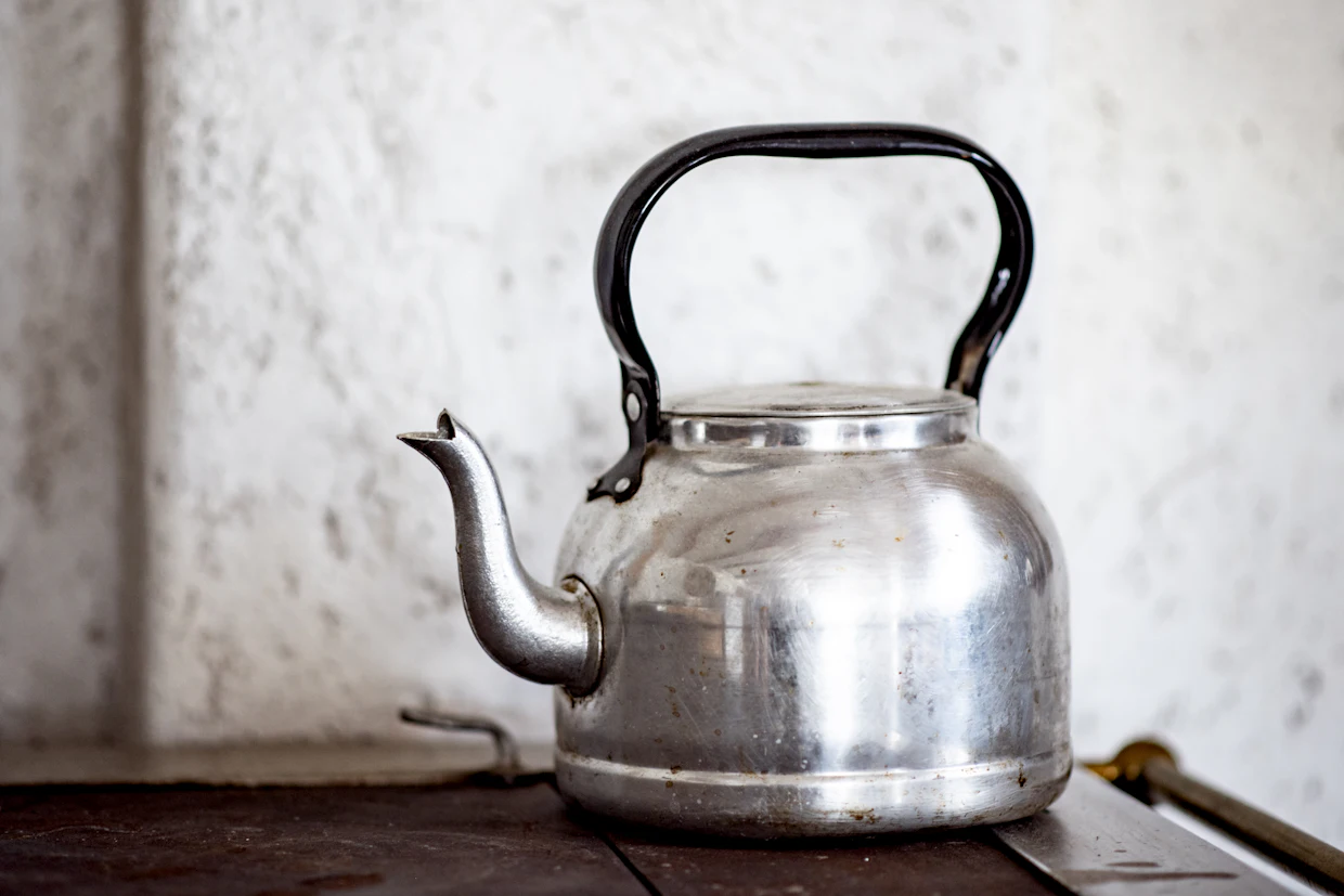A worn aluminum kettle sitting on a dark stove against a rough white wall.