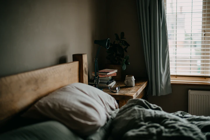 A mug on a wooden bedside table beside an unmade bed, with daylight through half-drawn curtains.
