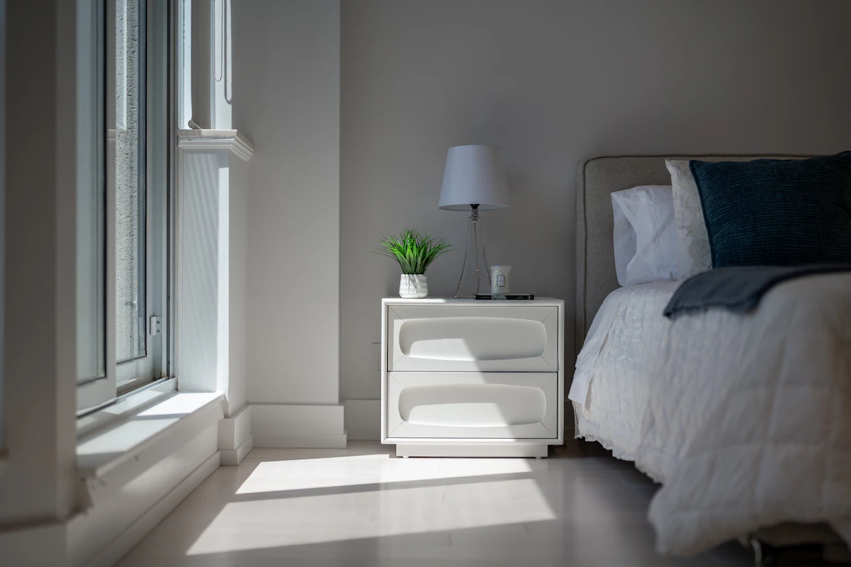 A bedroom in early morning light, with a nightstand beside a made bed.