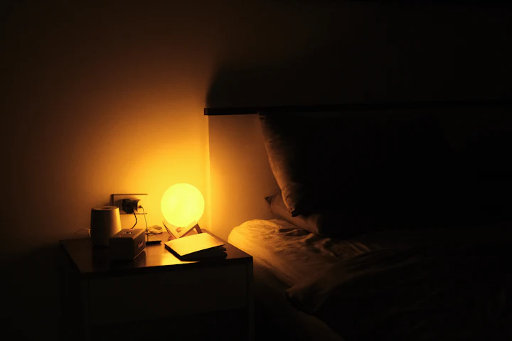 A nightstand at night, lit by a small lamp, beside a bed.