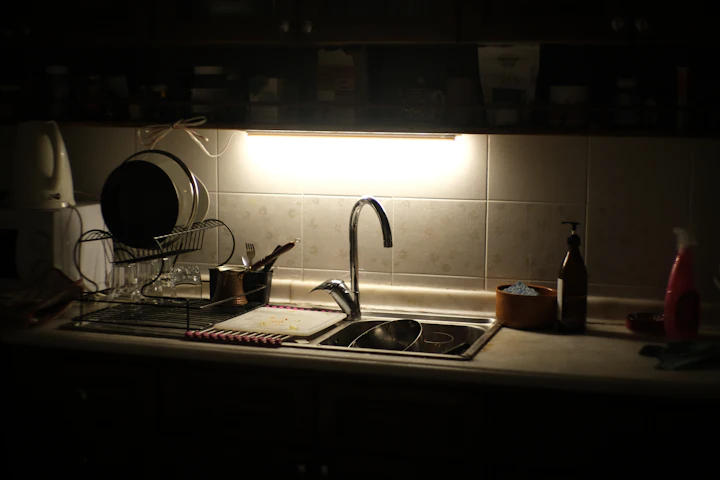 A kitchen counter at night lit by a single light over the sink, with dishes left in the rack.