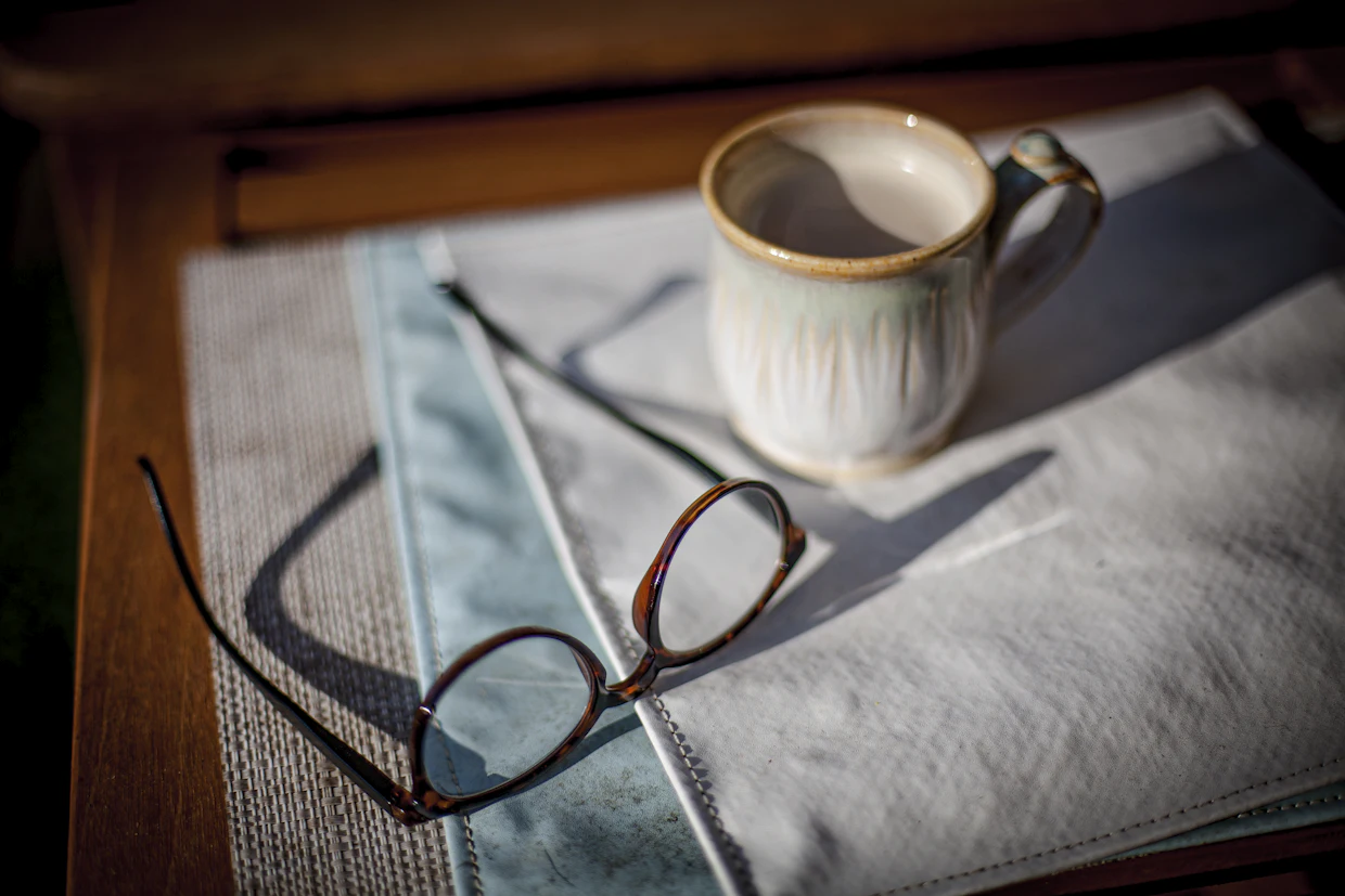 Une paire de lunettes de lecture et une tasse posées sur un napperon de lin, sur une table en bois, au soleil bas de l'après-midi.