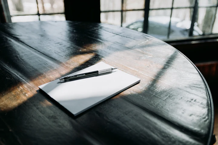 Un crayon posé sur un bloc de papier vierge sur une table ronde foncée près d'une fenêtre.