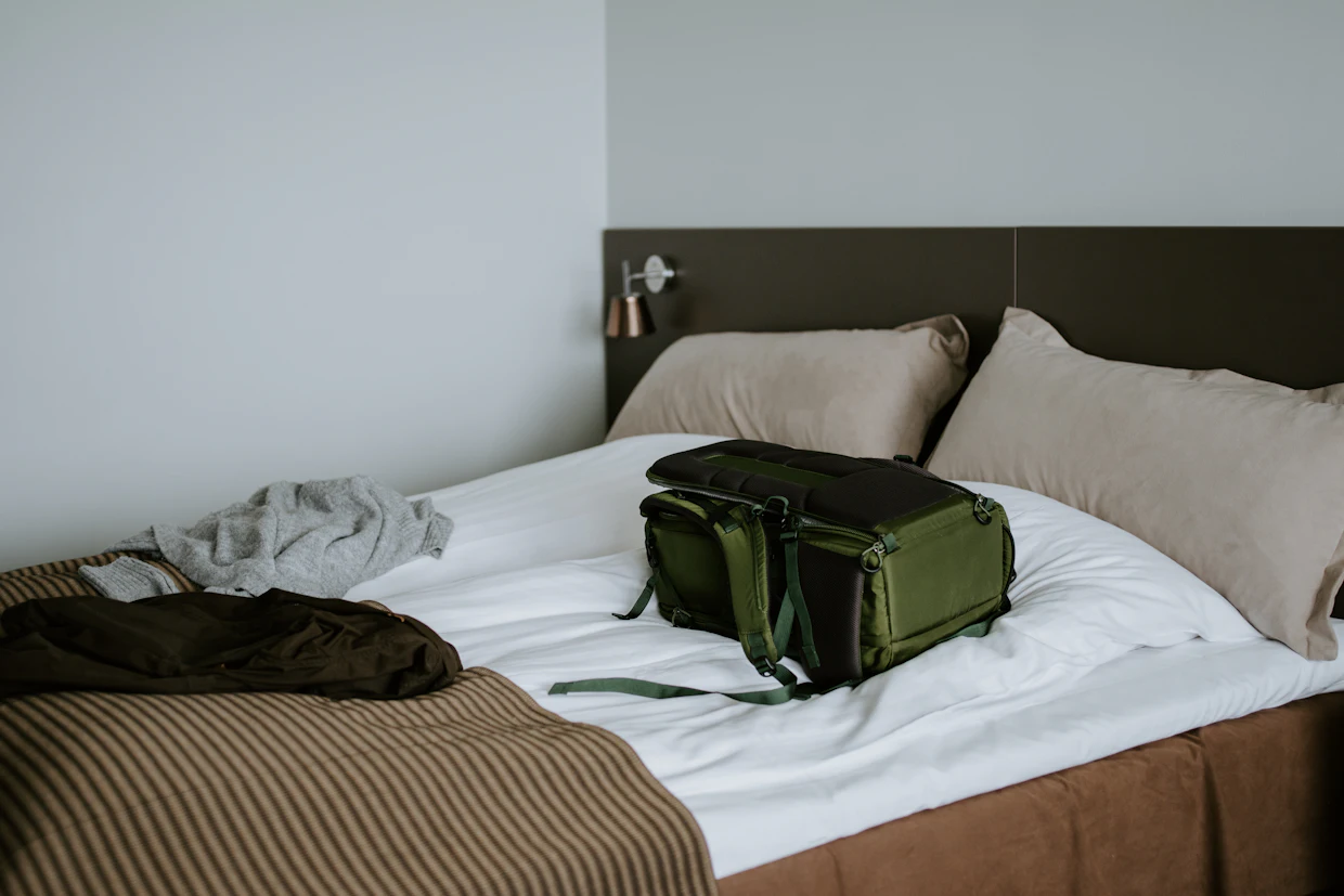 Un sac de voyage vert rempli, posé sur un lit fait, avec des vêtements pliés au coin du lit à côté.