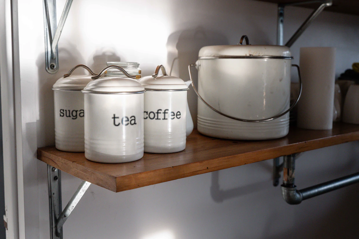 Trois boîtes de cuisine en émail étiquetées sucre, thé et café sur une tablette en bois.
