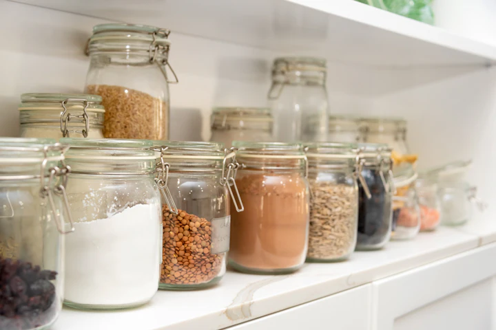 Une rangée de bocaux en verre à fermeture mécanique remplis de farine, de lentilles, d'avoine et de fruits séchés sur une tablette de cuisine.