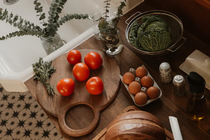 Des tomates sur une planche en bois, à côté d'une boîte d'œufs, d'un chou dans une passoire et d'un pain sur un comptoir de cuisine.