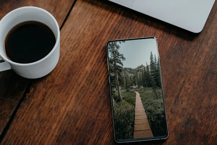 A phone lying face up on a wooden table beside a cup of black coffee.