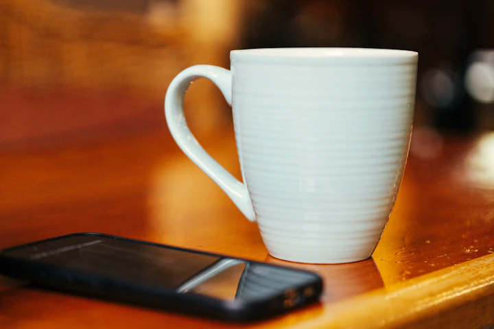 A phone lying beside a white mug on a wooden table.