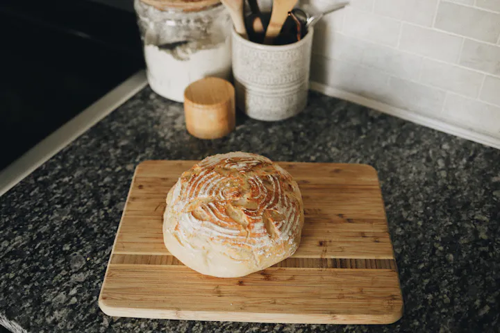 A round loaf of bread on a wooden board on a kitchen counter.