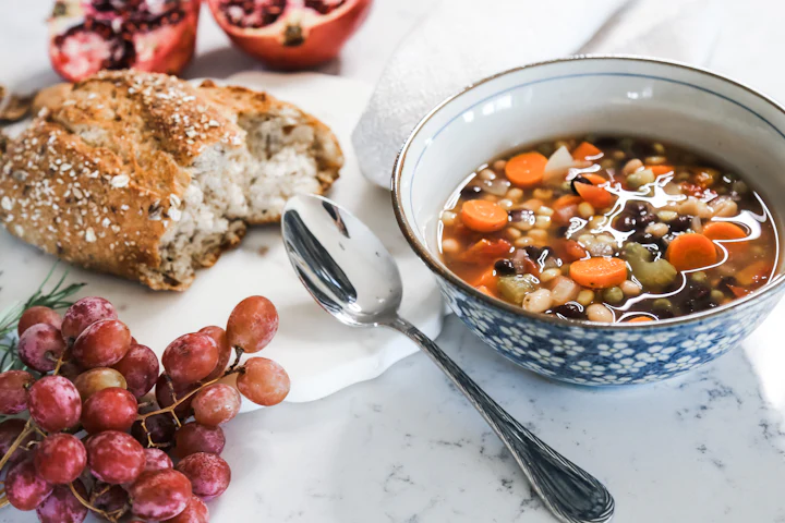 A bowl of vegetable soup with bread and grapes beside it.