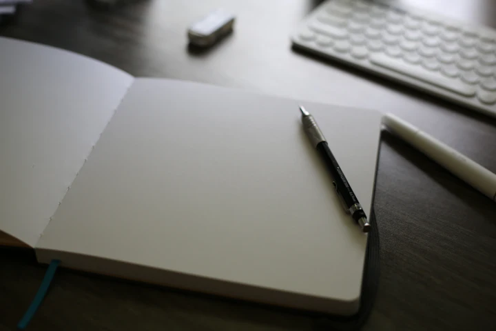 Un carnet ouvert aux pages vierges, avec un stylo posé en travers, sur un bureau en bois foncé.
